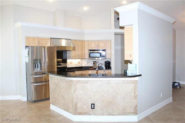 kitchen featuring dark countertops, backsplash, appliances with stainless steel finishes, light brown cabinets, and wall chimney range hood