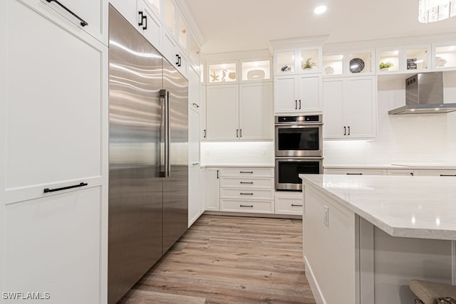 kitchen with light stone counters, light wood finished floors, stainless steel appliances, white cabinets, and wall chimney range hood