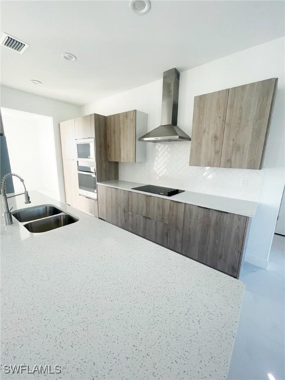 kitchen featuring modern cabinets, black electric stovetop, wall chimney range hood, stainless steel oven, and a sink