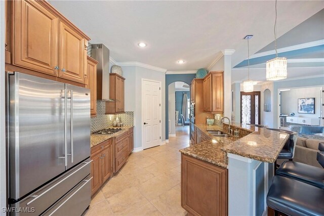 kitchen featuring arched walkways, stainless steel appliances, a sink, a kitchen breakfast bar, and wall chimney exhaust hood