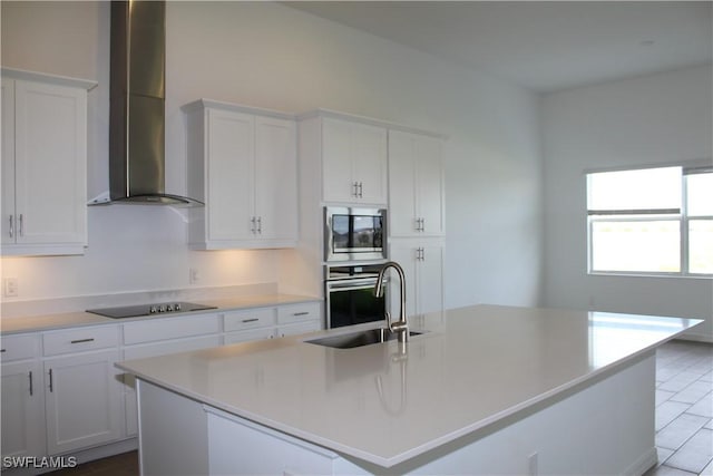 kitchen featuring a center island with sink, appliances with stainless steel finishes, white cabinets, a sink, and wall chimney range hood