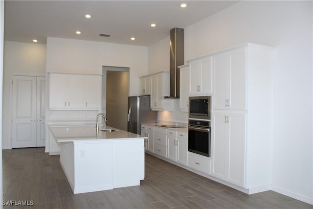 kitchen featuring a sink, white cabinets, appliances with stainless steel finishes, wall chimney range hood, and an island with sink