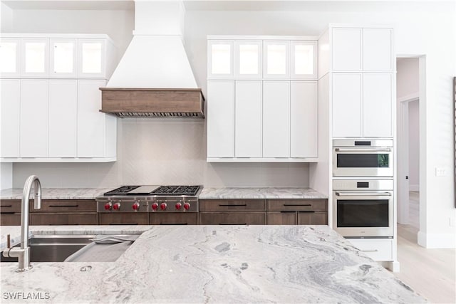 kitchen featuring white cabinetry, light stone counters, a sink, stainless steel appliances, and premium range hood