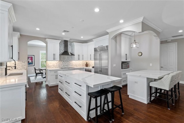 kitchen with arched walkways, a kitchen island, built in refrigerator, wall chimney range hood, and a sink