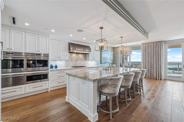 kitchen featuring a warming drawer, stainless steel appliances, backsplash, light wood-style flooring, and wall chimney exhaust hood