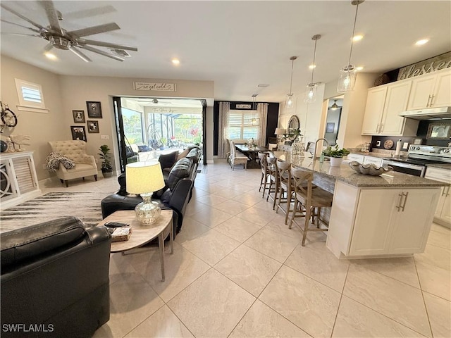 kitchen featuring under cabinet range hood, dark stone counters, open floor plan, stainless steel electric range oven, and an island with sink