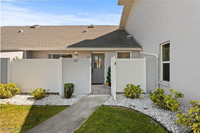 view of exterior entry with stucco siding, fence, and roof with shingles