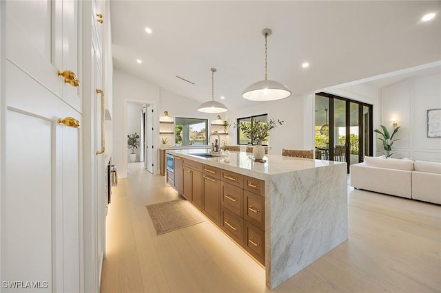 kitchen with pendant lighting, brown cabinets, a center island with sink, light wood-style flooring, and light stone countertops