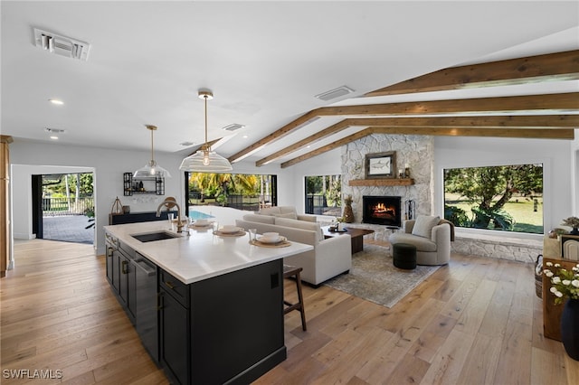 kitchen with a sink, lofted ceiling with beams, a stone fireplace, light wood-style flooring, and dark cabinets