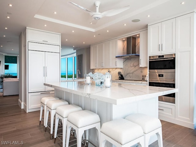 kitchen with wood finished floors, a tray ceiling, a kitchen island with sink, white cabinetry, and wall chimney range hood