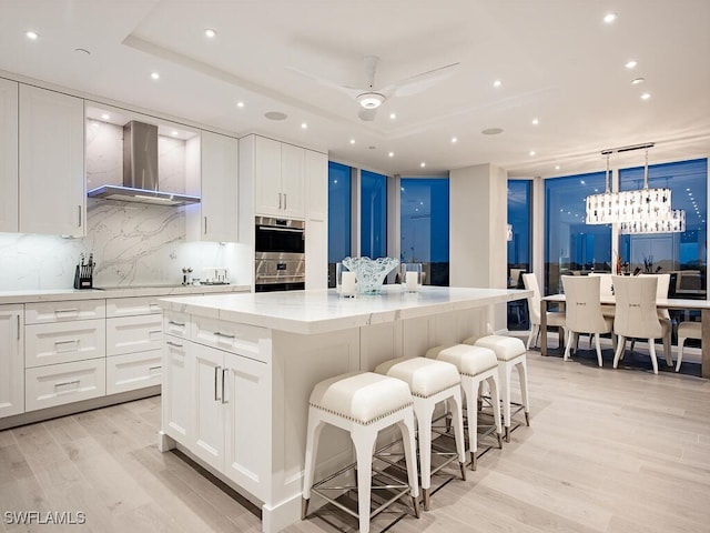 kitchen with a tray ceiling, light wood-style flooring, a large island, white cabinetry, and wall chimney exhaust hood