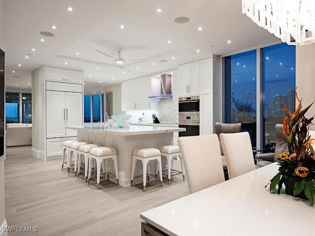 kitchen featuring paneled fridge, a large island with sink, white cabinetry, double oven, and wall chimney range hood