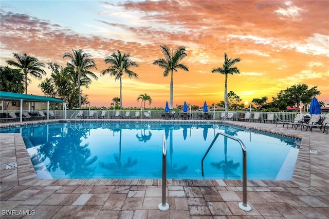 pool at dusk with a patio area, fence, and a community pool