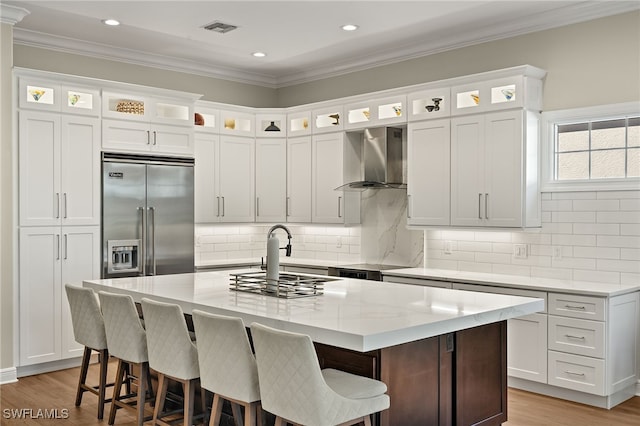 kitchen featuring wall chimney range hood, light wood-type flooring, ornamental molding, white cabinets, and built in fridge