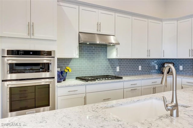 kitchen featuring decorative backsplash, appliances with stainless steel finishes, white cabinets, light stone countertops, and under cabinet range hood