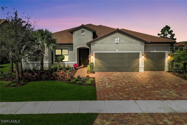 mediterranean / spanish-style home with an attached garage, a tiled roof, decorative driveway, a lawn, and stucco siding