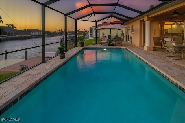 pool at dusk with glass enclosure, an outdoor pool, a patio area, and a water view