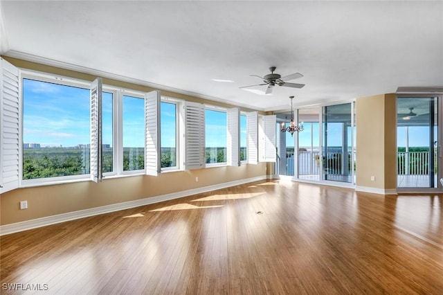 empty room featuring plenty of natural light, ceiling fan with notable chandelier, baseboards, and wood finished floors