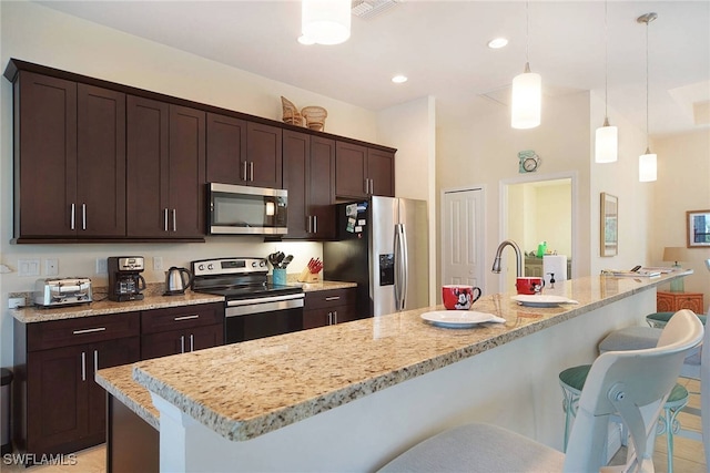 kitchen featuring dark brown cabinetry, appliances with stainless steel finishes, a breakfast bar area, light stone countertops, and pendant lighting