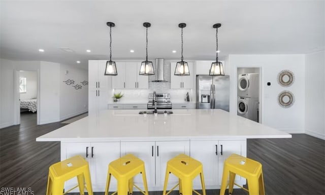 kitchen featuring stacked washer and clothes dryer, light countertops, appliances with stainless steel finishes, a large island with sink, and wall chimney exhaust hood