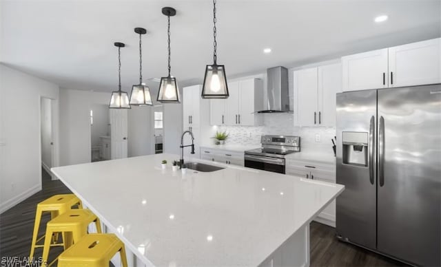 kitchen with appliances with stainless steel finishes, dark wood-type flooring, a kitchen island with sink, a sink, and wall chimney exhaust hood