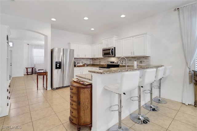 kitchen with arched walkways, light tile patterned floors, light stone counters, stainless steel appliances, and a peninsula