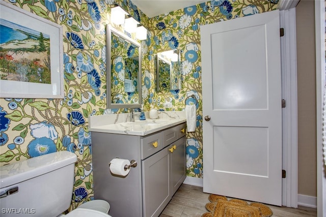 half bathroom featuring toilet, wallpapered walls, wood finished floors, and vanity