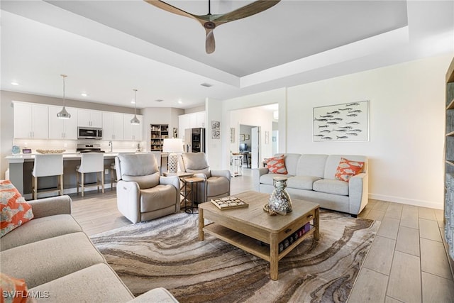 living room featuring baseboards, ceiling fan, a tray ceiling, light wood-type flooring, and recessed lighting