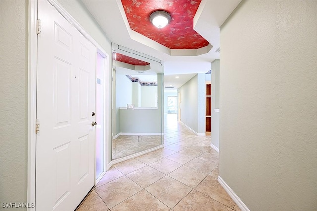 hall with a tray ceiling, light tile patterned floors, a textured wall, and baseboards