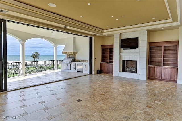 unfurnished living room with a water view, a fireplace, ornamental molding, a raised ceiling, and ornate columns