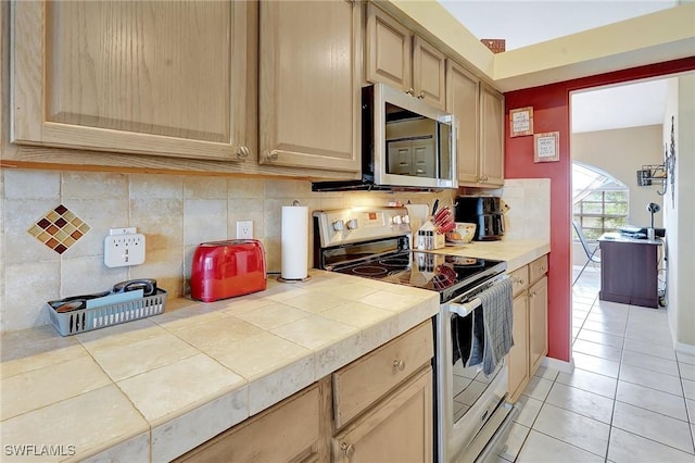 kitchen with light tile patterned floors, light brown cabinets, stainless steel appliances, tile counters, and backsplash