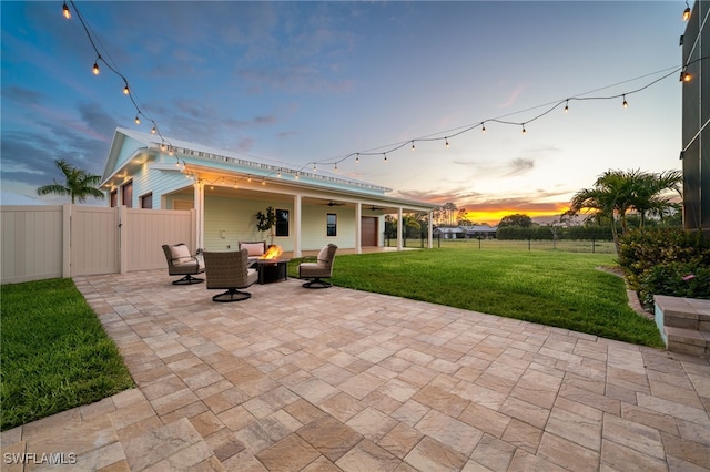 patio terrace at dusk with an outdoor fire pit, a gate, a fenced backyard, and a lawn