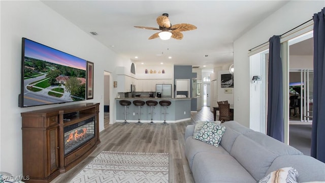 living room featuring light wood-style flooring, baseboards, ceiling fan, and a glass covered fireplace