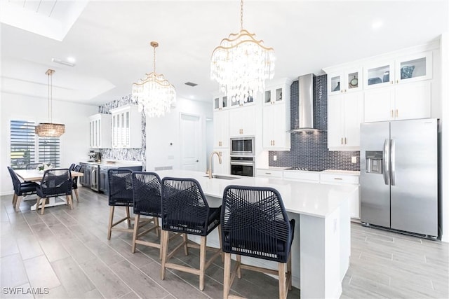 kitchen featuring appliances with stainless steel finishes, a kitchen island with sink, light countertops, wall chimney range hood, and a chandelier