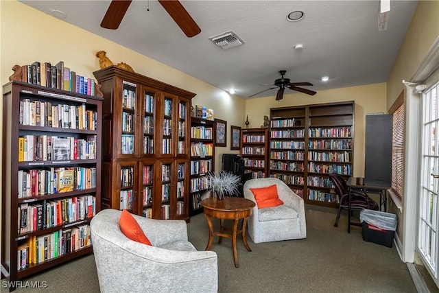 living area featuring ceiling fan, wall of books, carpet flooring, and visible vents