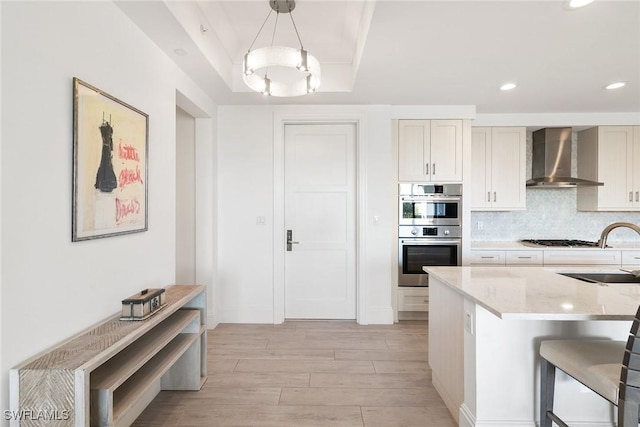 kitchen with a sink, tasteful backsplash, stainless steel double oven, wall chimney range hood, and a raised ceiling