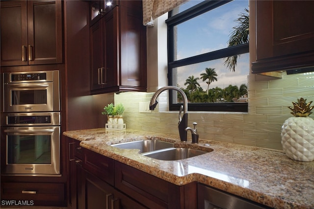 kitchen with light stone counters, decorative backsplash, double oven, glass insert cabinets, and a sink