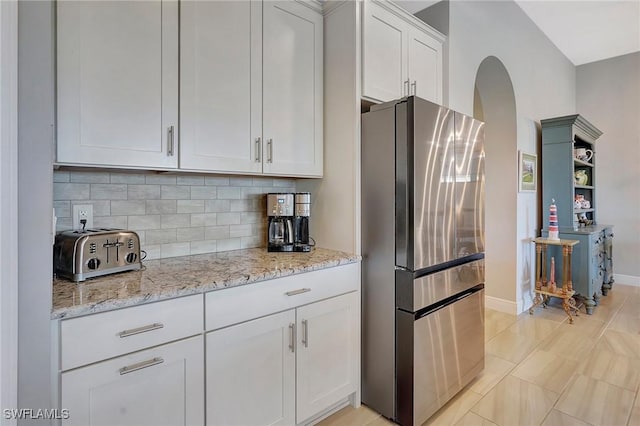 kitchen with freestanding refrigerator, arched walkways, white cabinetry, light stone counters, and backsplash