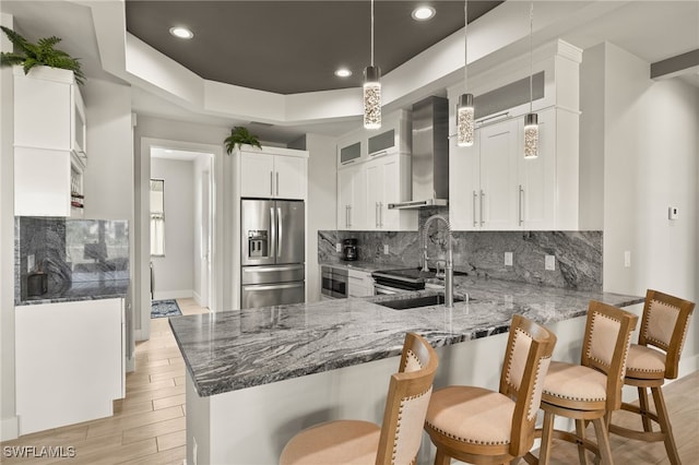 kitchen featuring stainless steel refrigerator with ice dispenser, a raised ceiling, stone countertops, wall chimney range hood, and a peninsula