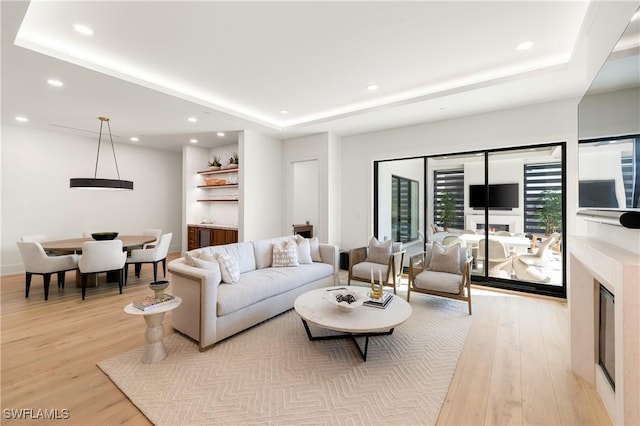 living room featuring light wood-style floors, recessed lighting, a raised ceiling, and a glass covered fireplace