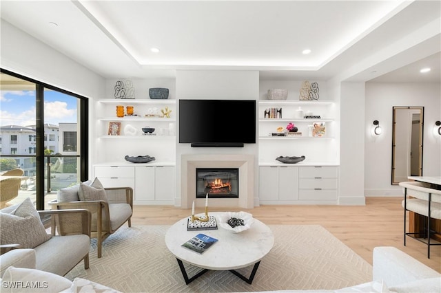 living area with recessed lighting, light wood-style flooring, a tray ceiling, and a glass covered fireplace