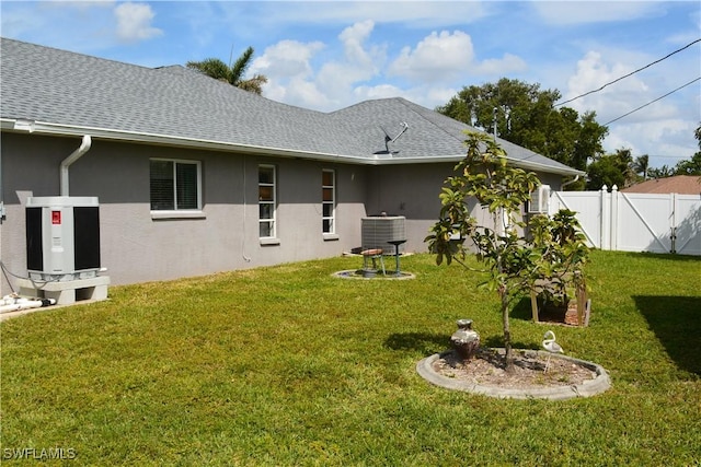 rear view of property with a shingled roof, a lawn, fence, central AC, and stucco siding