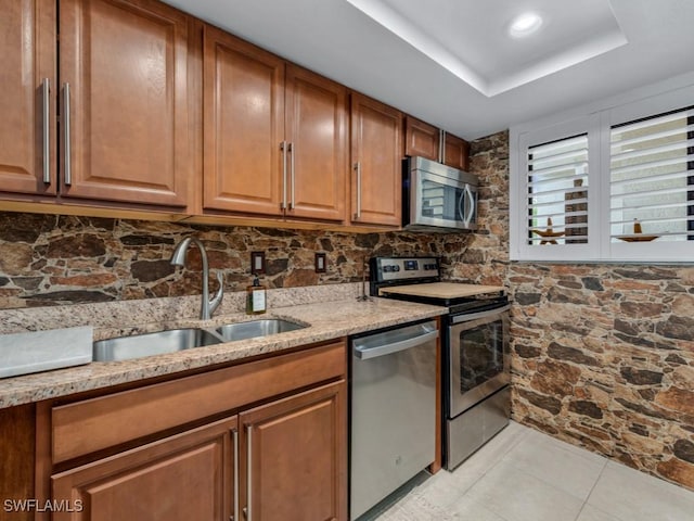 kitchen featuring light stone counters, stainless steel appliances, a sink, brown cabinets, and a raised ceiling