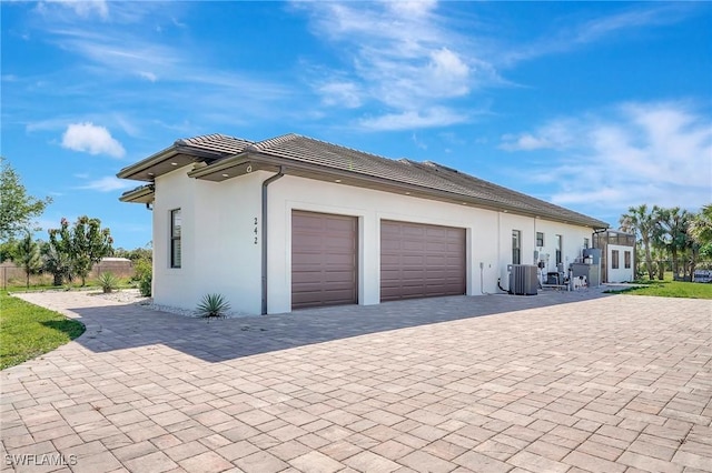 view of property exterior with stucco siding, a tile roof, decorative driveway, central AC, and a garage