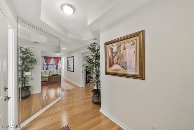 corridor featuring baseboards, a raised ceiling, and light wood-style flooring