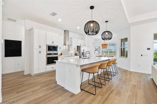 kitchen with stainless steel appliances, backsplash, light wood-style flooring, wall chimney range hood, and a large island with sink