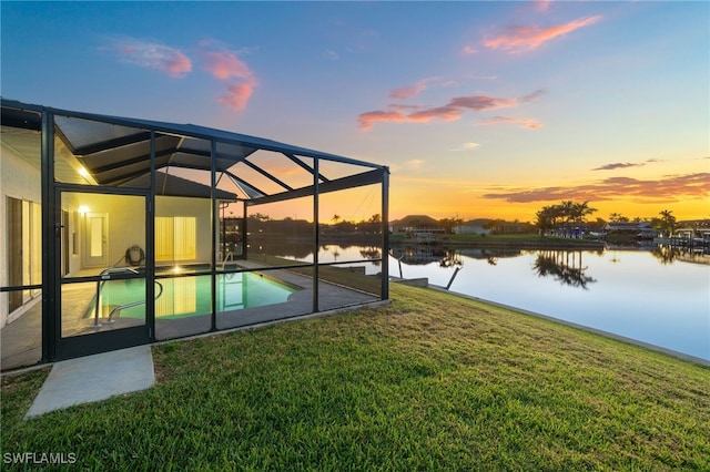 pool at dusk with an outdoor pool, glass enclosure, a water view, a yard, and a patio area