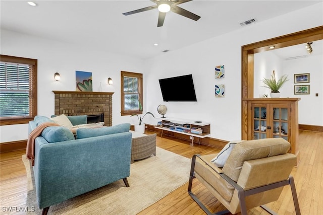 living room with a ceiling fan, visible vents, baseboards, a brick fireplace, and light wood finished floors