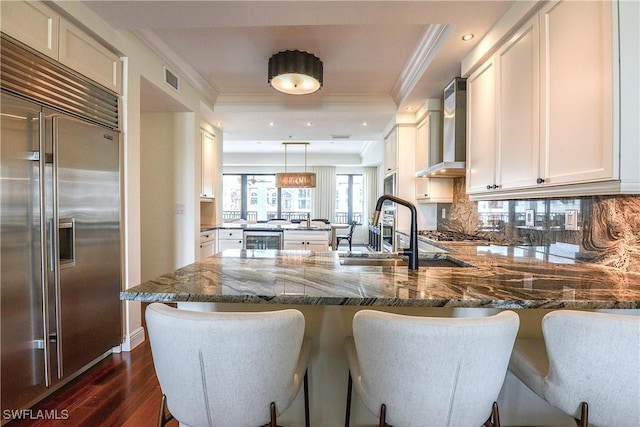 kitchen featuring beverage cooler, a peninsula, wall chimney range hood, stainless steel built in fridge, and dark stone countertops