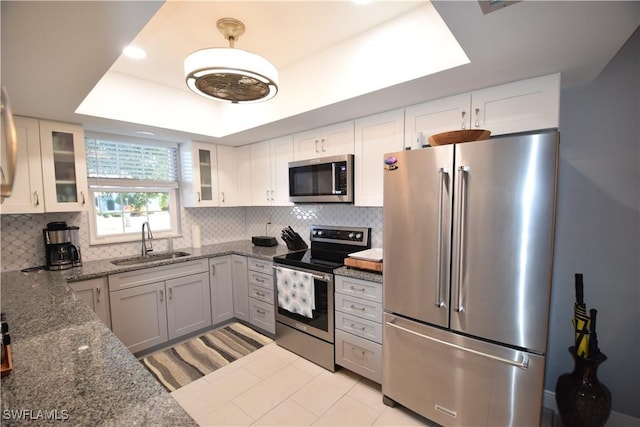 kitchen featuring dark stone counters, a tray ceiling, a sink, decorative backsplash, and stainless steel appliances